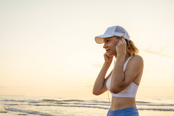 Euroopan näköinen tyttö urheiluvaatteissa kuuntelee musiikkia kuulokkeilla meren rannalla aamulenkillä. Laadukas kuva - Valokuva, kuva