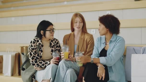 Średnie ujęcie trzech młodych wielonarodowych dziewczyn z plastikowymi kubkami na wynos z napojami gazowanymi siedzącymi na ławce w centrum handlowym i rozmawiającymi - Materiał filmowy, wideo