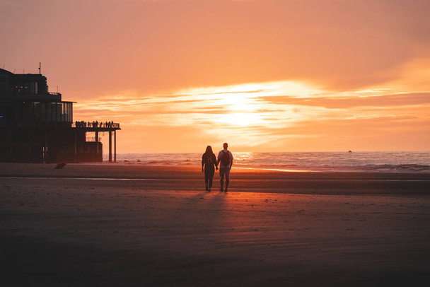 日没のベルギー西部のオストエンデのビーチで若いカップルのロマンチックな散歩. 愛と献身。 水のプールでの反射. - 写真・画像