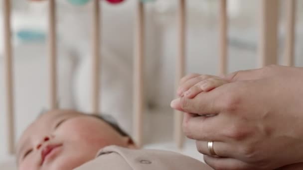 Zbliżenie nierozpoznawalnej kobiety trzymającej malutką rączkę syna śpiącego spokojnie w łóżeczku w dzień - Materiał filmowy, wideo