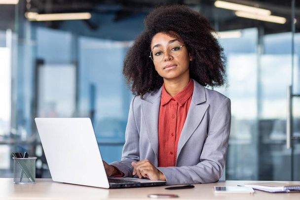Portrait of a serious and successful African American businesswoman sitting in the office at the desk, working on a laptop and looking confidently at the camera. - 写真・画像