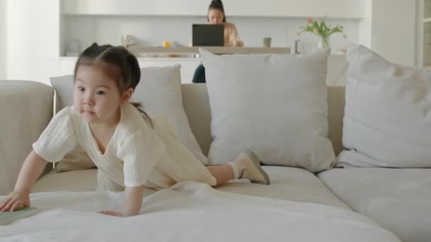 Plan moyen de gentille petite fille asiatique prenant soin de son lièvre doux préféré le couvrant tendrement avec une couverture tout en jouant à la maison. Sa mère travaille sur un ordinateur portable en arrière-plan - Séquence, vidéo