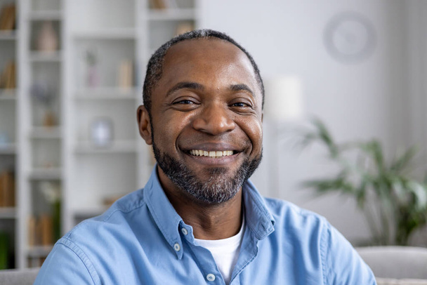 Close-up retrato de maduro experiente afro-americano homem sorrindo e olhando para a câmera enquanto sentado no sofá na sala de estar em casa. - Foto, Imagem