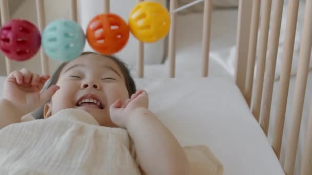 Medium close-up van mooi aziatisch klein meisje hebben plezier tijdens het spelen met kleurrijke rammelaars opknoping op haar wieg - Video