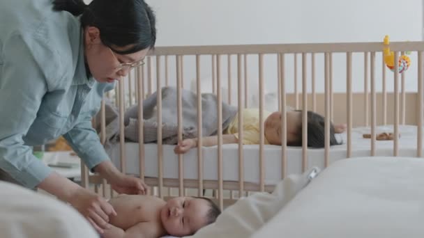 Střední záběr mladé asijské ženy posilující svaly nohou svého novorozeného syna tím, že fyzické cvičení a masírovat ho ležící na zádech v postýlce. Asijské batole dívka spací v pozadí - Záběry, video