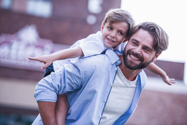 Handsome bearded dad and his cute little son are smiling while playing together outside, boy is sitting piggyback - Photo, Image