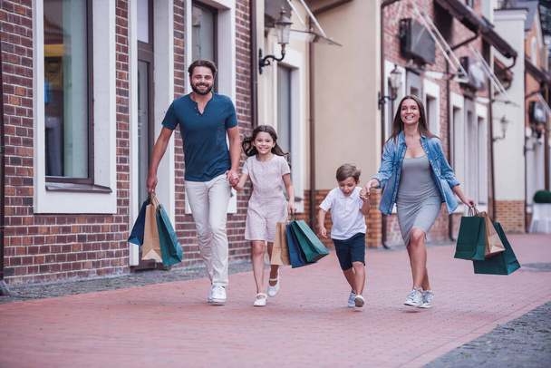 Hermosos padres y sus lindos niños pequeños están llevando bolsas de compras, tomados de la mano y sonriendo mientras caminan por la calle
 - Foto, imagen