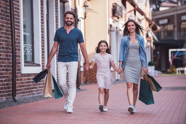 Beautiful parents and their cute little daughter are carrying shopping bags, holding hands and smiling while walking down the street - Photo, Image