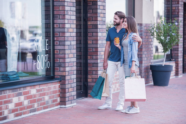 Mooie man en vrouw zijn uitvoering van boodschappentassen, etalage kijken en glimlachen tijdens het wandelen in de straat - Foto, afbeelding