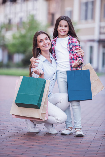 Beautiful mom and her cute little daughter are holding shopping bags, looking at camera and smiling while standing outdoors - Photo, Image
