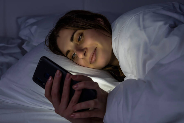 Una ragazza a letto con un telefono su un letto bianco in una stanza buia - Foto, immagini