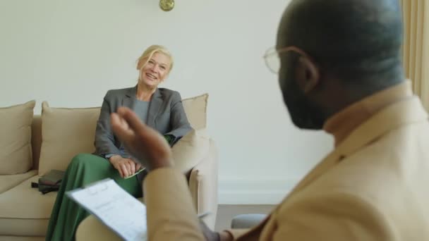 Olkapään yli laukaus kypsä nainen istuu sohvalla, kuunnellen neuvoja Afrikkalainen amerikkalainen terapeutti ja hymyillen aikana neuvonta istunto - Materiaali, video