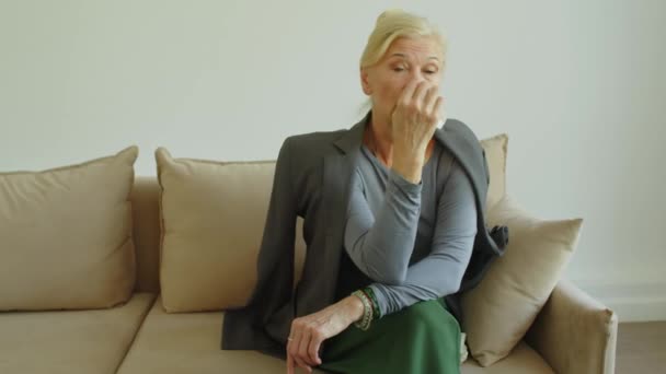 Medium long shot of upset senior woman wiping her tears with napkin and posing for camera while sitting on sofa in therapist office - Кадры, видео