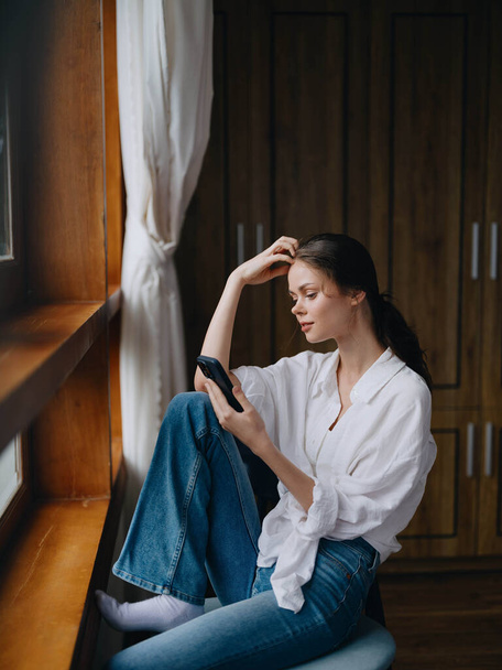 Young woman sitting at the window on a chair with a phone in hand smiling and looking out the window, spring mood, home cozy atmosphere, aesthetic lifestyle. High quality photo - Photo, Image