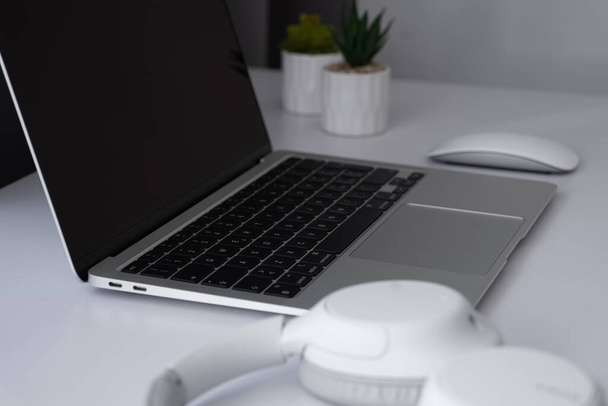 Zijaanzicht op een lege scherm laptop met muis, koptelefoon en planten op een witte tafel thuis kantoor. - Foto, afbeelding