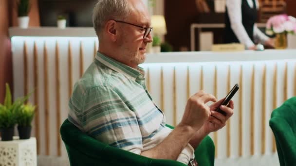 Rentner checkt Smartphone-App im Lounge-Bereich eines Fünf-Sterne-Hotels und navigiert mit Online-Browser in den sozialen Medien. Ältere Reisende entspannen sich in der Lobby des Ferienhauses. Handschuss. - Filmmaterial, Video