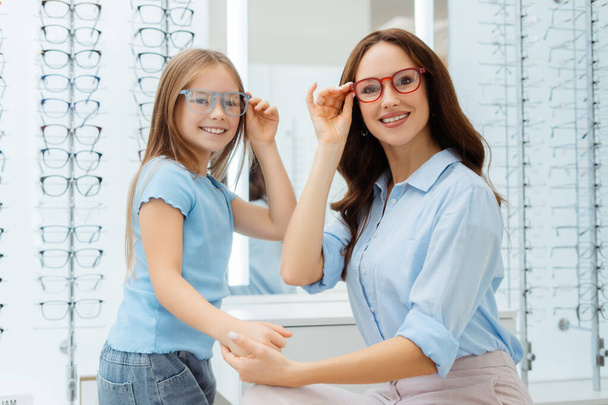 Glimlachende moeder en haar dochter proberen een bril op te doen bij de optiek. Gezondheidszorg, gezinsconcept - Foto, afbeelding