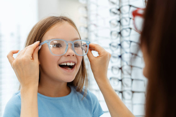 Jovem mãe ajudando sua filha a escolher óculos na loja óptica. Saúde, conceito de família - Foto, Imagem