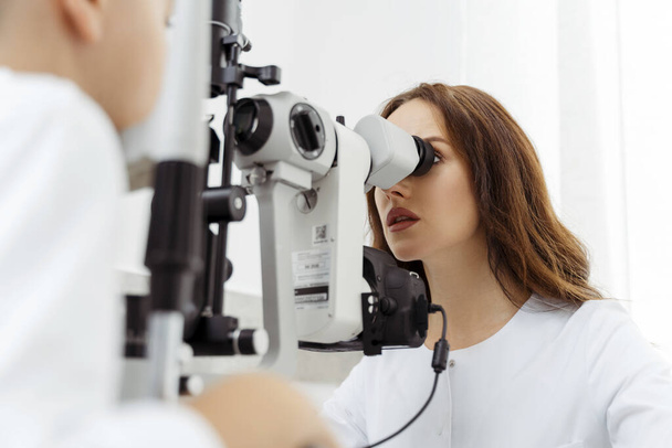 Optometrist in whitecoat kijkend door lens van gezichtscontrole apparatuur in het bijzijn van kleine jongen. Gezondheidszorgconcept - Foto, afbeelding