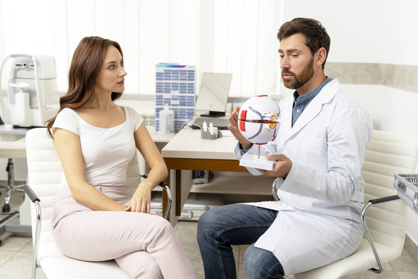 ophtalmologiste masculin utilisant un modèle de globe oculaire et parlant avec une patiente à la clinique. Soins de santé, concept scientifique - Photo, image
