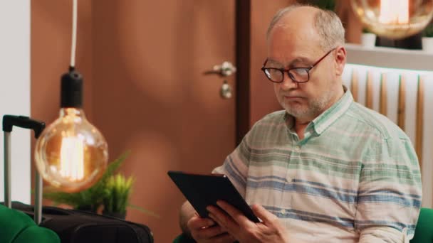 Older man tourist on lounge area sofa, surfing the web on digital tablet. Person holding device to browse internet waiting to see room at five star hotel on vacation, checks online reservation. - Footage, Video