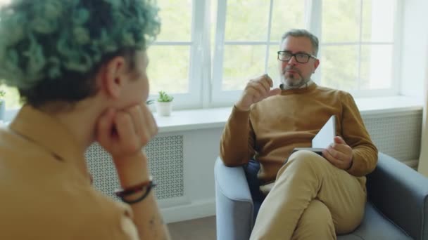 Přes rameno snímek dospělé mužské psycholog s šedými vlasy mluvit s dívkou s modrými vlasy během konzultace v psychiatrické léčebně - Záběry, video
