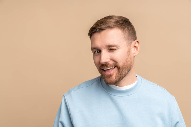 Flirting man winks look at camera, studio portrait isolated on beige. Bearded middle aged guy smiling winking playfully jokingly impishly with one eye, hinting, hidden intent, non-verbal communication - Photo, Image