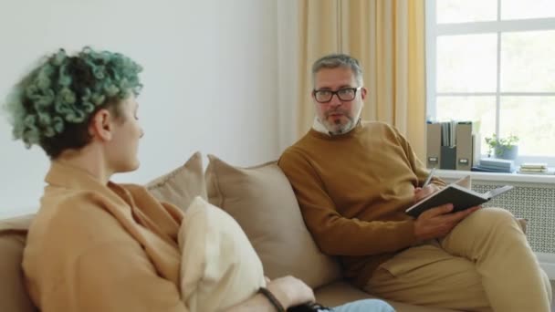 Starší mužský terapeut sedí na pohovce, čte si poznámky a radí se s mladou pacientkou - Záběry, video