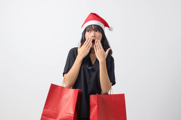Portrait of surprised Asian woman with red Santa hat feeling happy while purchasing stuff. Excited girl holding shopping bag. New year and christmas concept. Isolated image on white background - Photo, Image