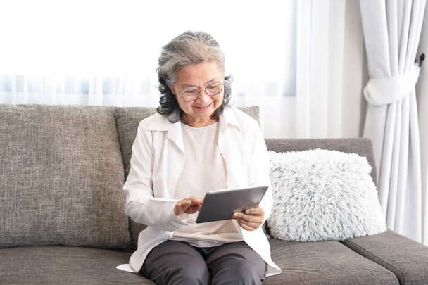 asiatica anziana seduta sul divano, rilassarsi in soggiorno, utilizzando internet online su tablet per comunicare con la sua famiglia, concetto di anziani vita moderna sulla tecnologia - Foto, immagini