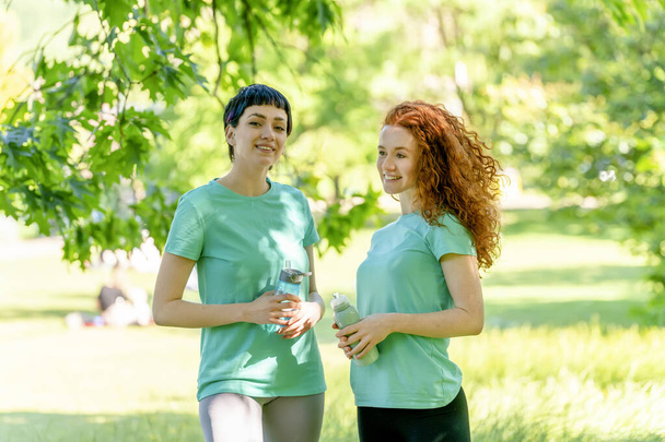 Gelukkige vrouwen rusten en drinken water na het sporten in het park. Fitness, sport en lichaamsbeweging - Foto, afbeelding