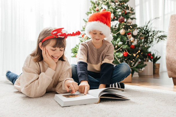 Happy children, a boy and a girl, read a book on Christmas or New Years Eve against the background of a Christmas tree. Waiting for the holidays. - Photo, Image