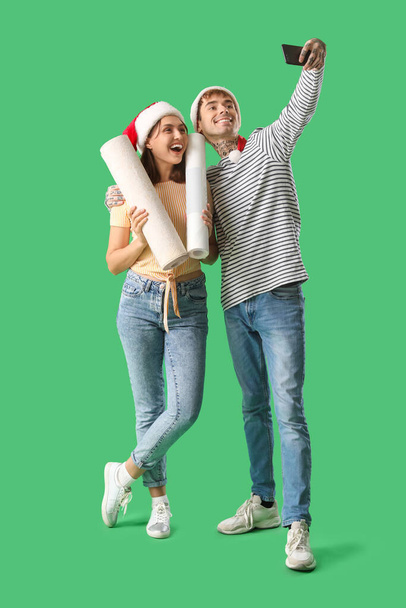 Young couple in Santa hats with wallpapers taking selfie on green background - Photo, Image