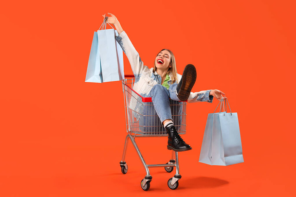 Beautiful young stylish woman with shopping bags sitting in trolley on orange background - Photo, Image