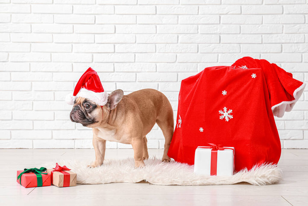 Niedliche Französische Bulldogge mit Weihnachtsmütze und Tasche voller Weihnachtsgeschenke in der Nähe weißer Ziegelmauer - Foto, Bild