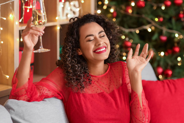 Gyönyörű afro-amerikai nő pezsgővel otthon a kanapén. Újévi ünneplés - Fotó, kép