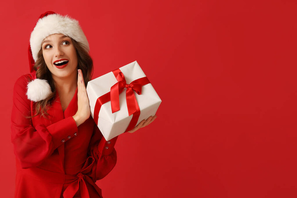 Surprised young woman in Santa hat with Christmas gift box on red background - Photo, Image
