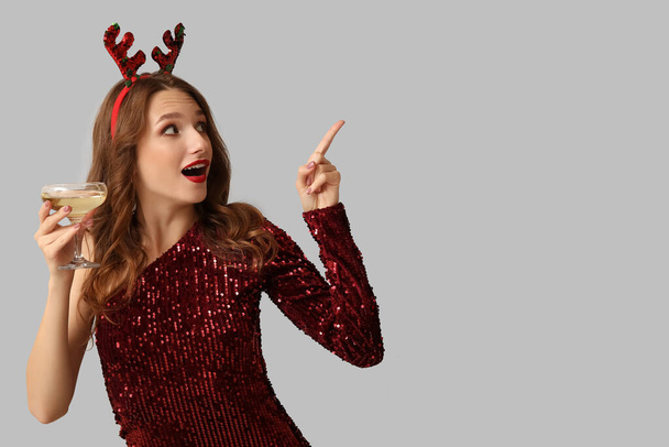 Young woman in reindeer horns holding glass of champagne and pointing at something on grey background - Photo, Image