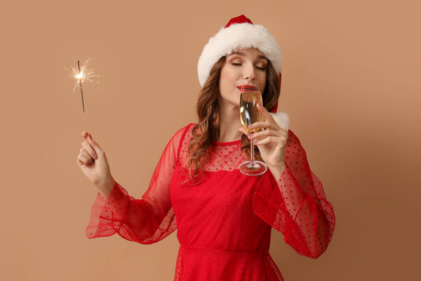 Junge Frau mit Weihnachtsmütze mit Wunderkerze und Glas Champagner auf braunem Hintergrund - Foto, Bild