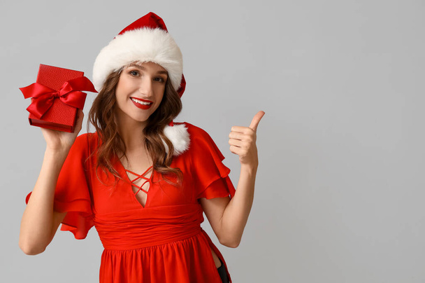 Молодая женщина в шляпе Санты держит рождественскую подарочную коробку и показывает большой палец вверх жест на сером фоне - Фото, изображение