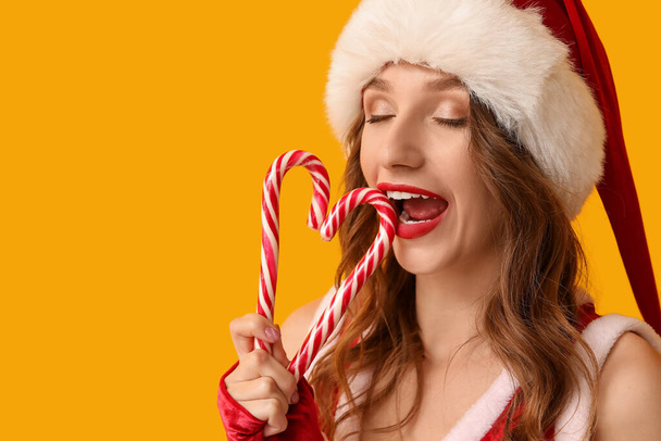 黄色の背景にクリスマスキャンディー缶とサンタクロースのコスチュームの若い女性 - 写真・画像