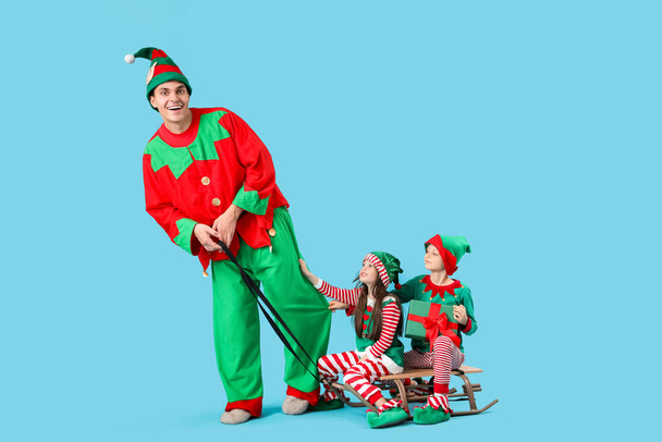 Father and cute little children in elf's costumes with gift box on sledge against blue background - Photo, Image