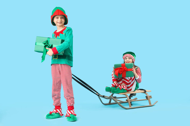 Cute little children in elf's costumes with gift boxes and sledge on blue background - Fotó, kép
