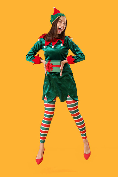 Beautiful young woman in elf's costume with gift box jumping on yellow background - Photo, Image