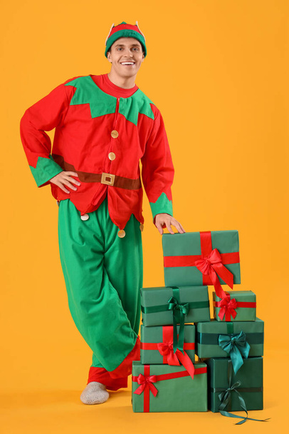 Handsome young man in elf's costume with gift boxes on yellow background - Photo, Image