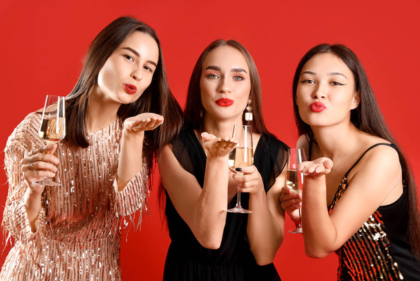 Όμορφες νεαρές γυναίκες με σαμπάνια να φυσάει φιλιά σε κόκκινο φόντο. Πρωτοχρονιά εορτασμού - Φωτογραφία, εικόνα