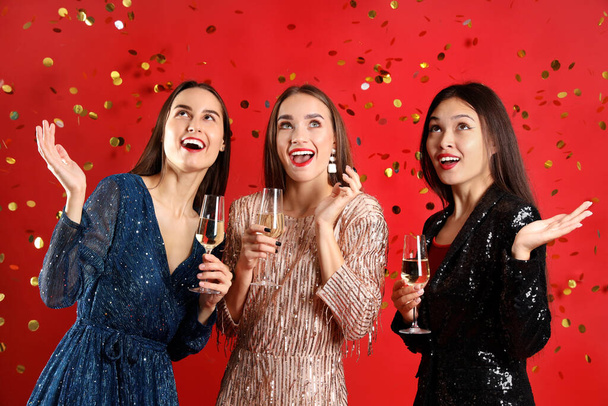 Gyönyörű fiatal nők pezsgővel és konfettivel vörös háttérrel. Újévi ünneplés - Fotó, kép