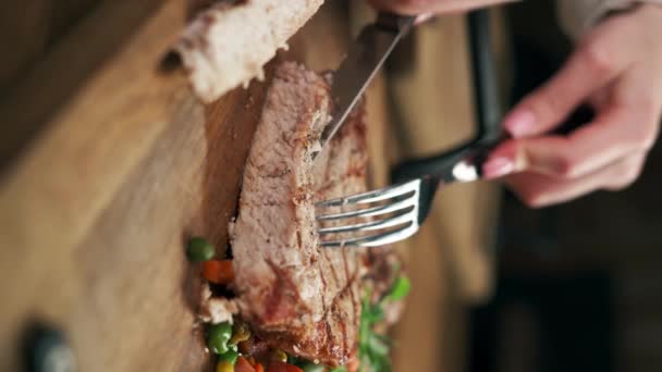 Frau schneidet mit Messer und Gabel gegrilltes Rindersteak auf Holzbrett. Grillfleisch zum Abendessen im Restaurant. Vertikales Video - Filmmaterial, Video