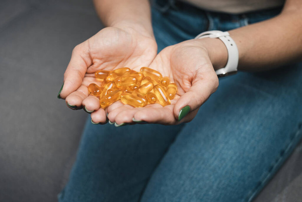 Mulher jovem segura pilha de ómega-3 suplementos de óleo de peixe, cápsulas de vitamina amarela em suas mãos, sentado no sofá em casa, vista de close-up - Foto, Imagem