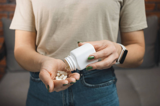 Mujer joven sostiene cápsulas médicas blancas y pastillas en una mano, tomar medicamentos de un frasco, vista de cerca. - Foto, Imagen